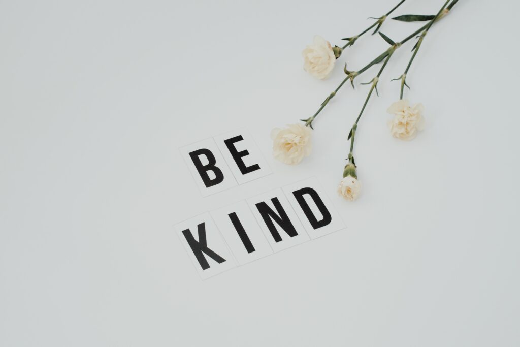 Minimalist image of 'Be Kind' text with white flowers on a light background, promoting kindness.