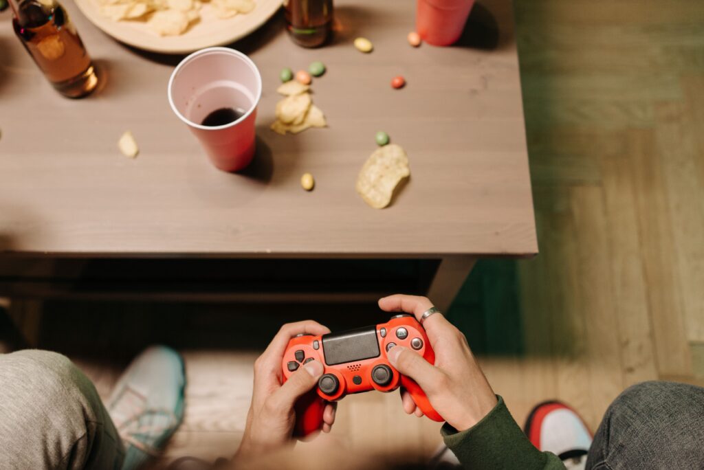 Person holding a red game controller with snacks and drinks around, top view.