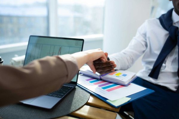 Two business professionals shaking hands over graphs and reports in an office setting.